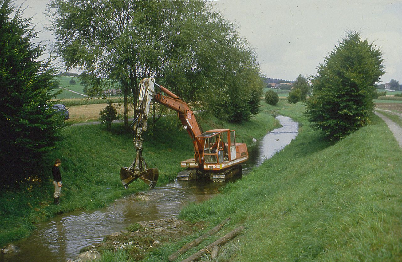 Ein Bagger steht in einem Bach, welcher von Feldern umgeben ist.