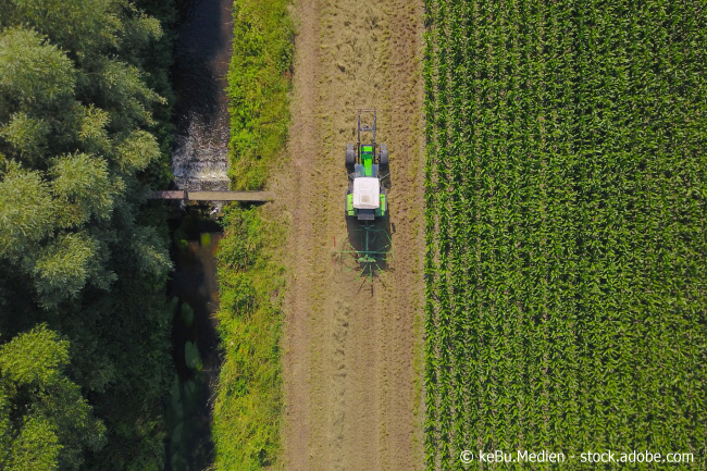 Luftbild von geradem Bach, direkt daneben ein Feld mit Nutzfahrzeig
