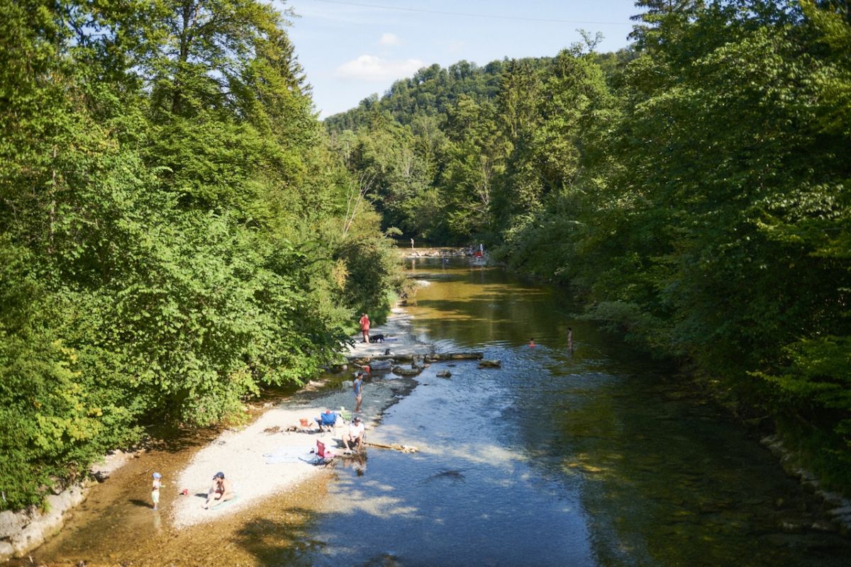 Blick auf einen kleinen Fluss mit Kiesbank, wo Menschen spielen und entspannen, der Fluss ist von Bäumen umgeben.